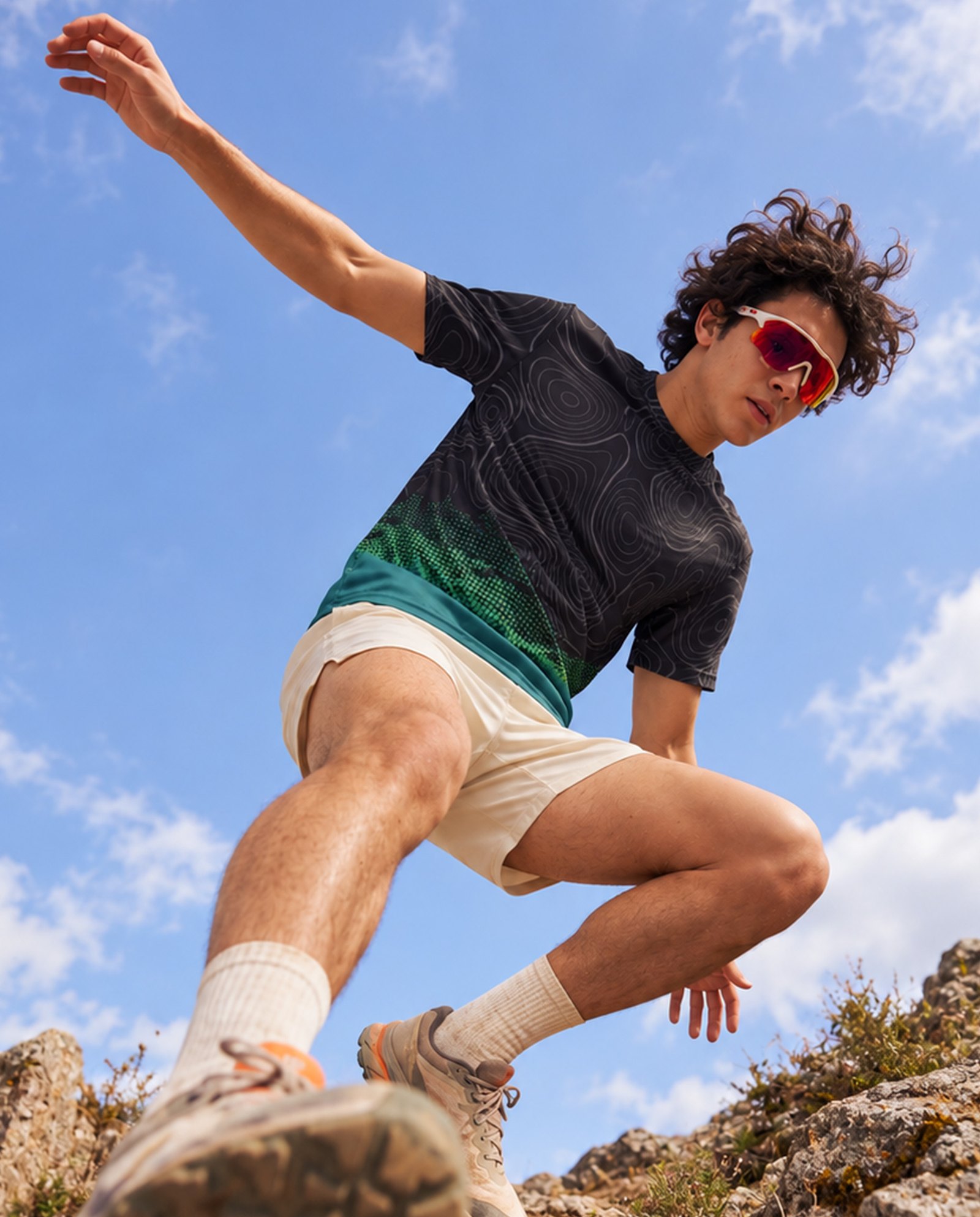 Art-directed AI photoshoot of a trail runner in a topo print performance tee leaping over a rocky ridge, shot from a low angle against blue sky
